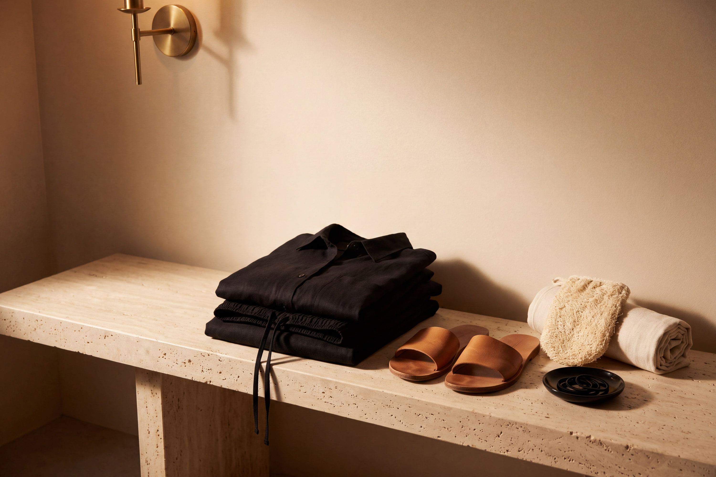 A loose black linen outfit, leather slides, and an exfoliating mitt laid out on a travertine bench
