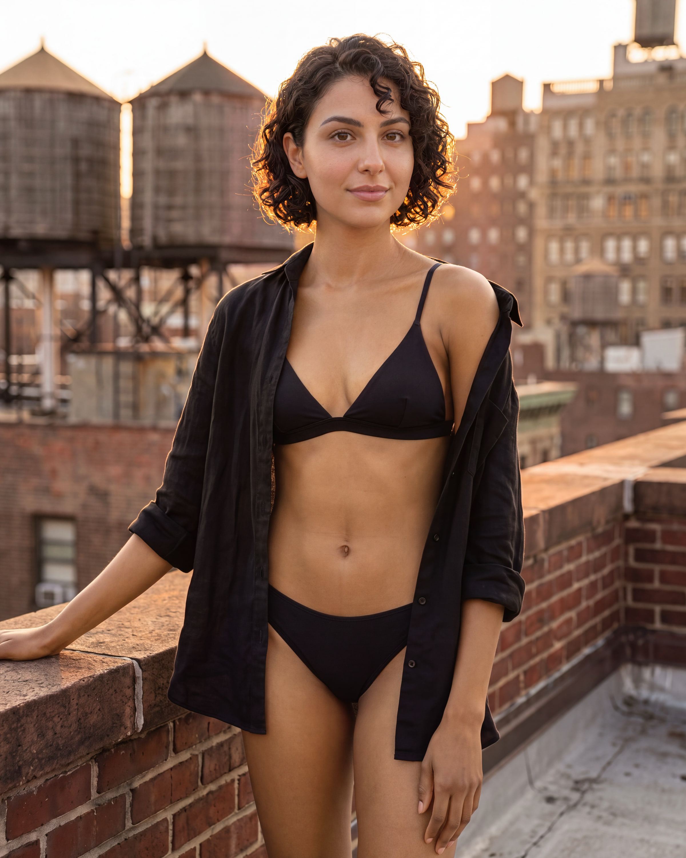 Woman with a golden custom spray tan on a city rooftop at golden hour, water towers soft behind her